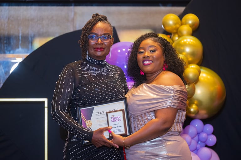 Two women in elegant attire holding awards at an event, posing in front of purple and gold balloon decorations