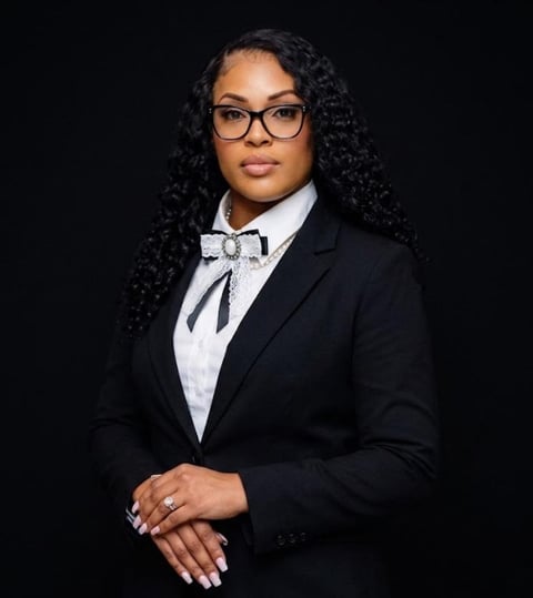 Professional portrait of a woman wearing a black blazer, white dress shirt, and patterned bow tie with glasses against a black background