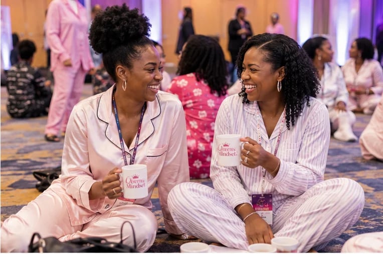 Two women in pajamas sitting on floor at pajama party event, holding drinks and conversing, with other guests visible in background