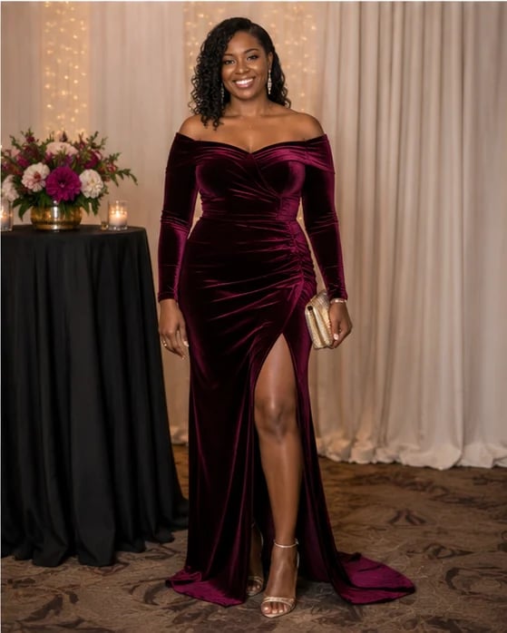 Woman in elegant burgundy velvet off-shoulder gown with high slit, standing in formal setting with floral arrangement and gold lighting